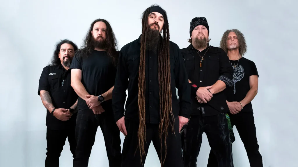 Five musicians in dark clothing stand against a white background, with the center figure prominently displaying long dreadlocks.