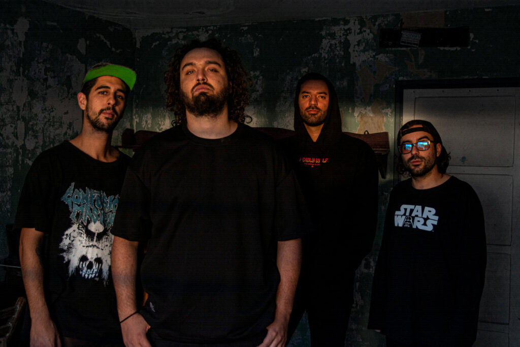 Four musicians wearing black band and graphic t-shirts stand against a weathered dark wall, with the leftmost wearing a neon green cap and the rightmost wearing blue-tinted sunglasses.